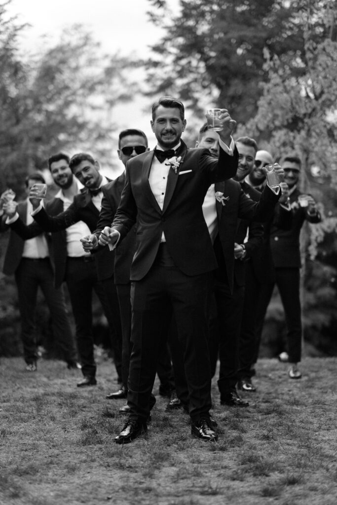 Un grupo de best menvestidos con trajes y corbatas formales posan para una foto en blanco y negro. Los árboles de fondo sugieren un entorno al aire libre. El novio en el frente sostiene una copa, buscando proponer un brindis.