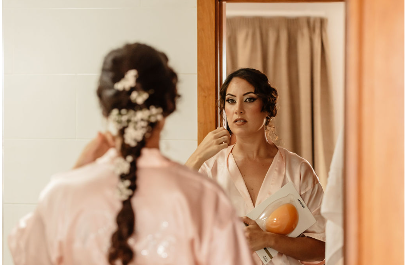 Persona con bata rosa claro y trenza adornada con flores blancas, mirándose en el espejo mientras se prepara para su boda. Sostiene un accesorio naranja en su mano izquierda.