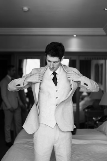 Hombre ajustándose su chaqueta de traje claro en una habitación de hotel, preparándose para su boda, con otra persona en traje visible en el fondo.