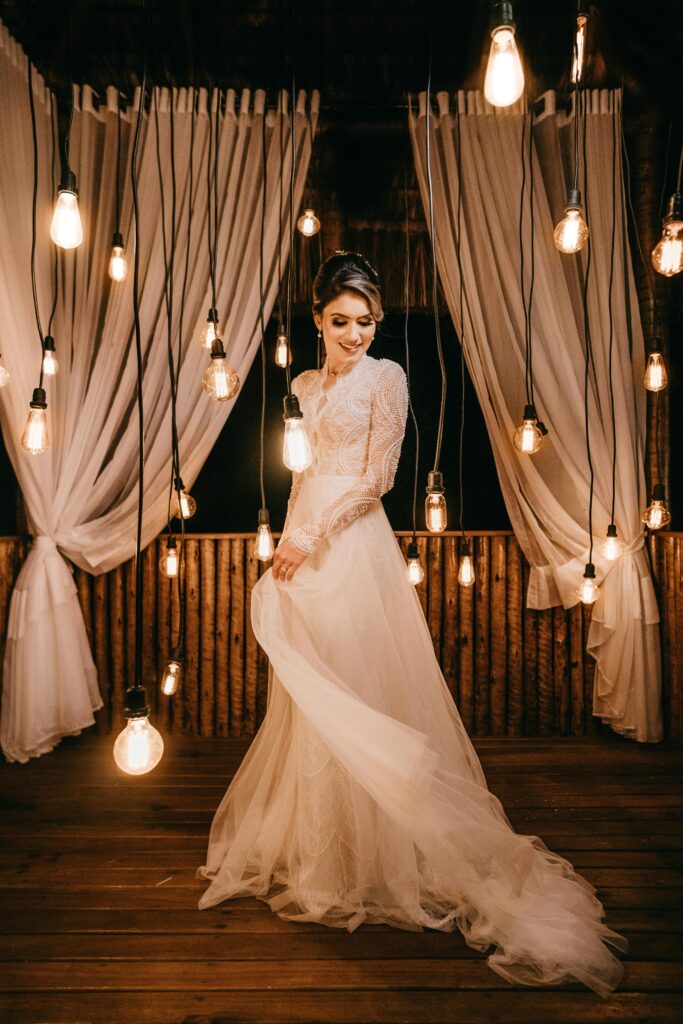 Persona de pie en un piso de madera, luciendo un elegante vestido blanco largo con detalles intrincados de encaje en el corpiño y las mangas. El fondo incluye cortinas blancas drapeadas y una pared de madera