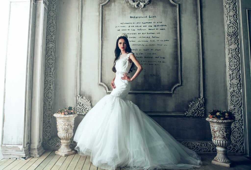 Persona vistiendo un elegante vestido de novia blanco posando frente a una pared decorativa de estilo vintage con intrincados diseños y dos jarrones con flores a cada lado. La persona está con una mano en la cadera y el otro brazo relajado.