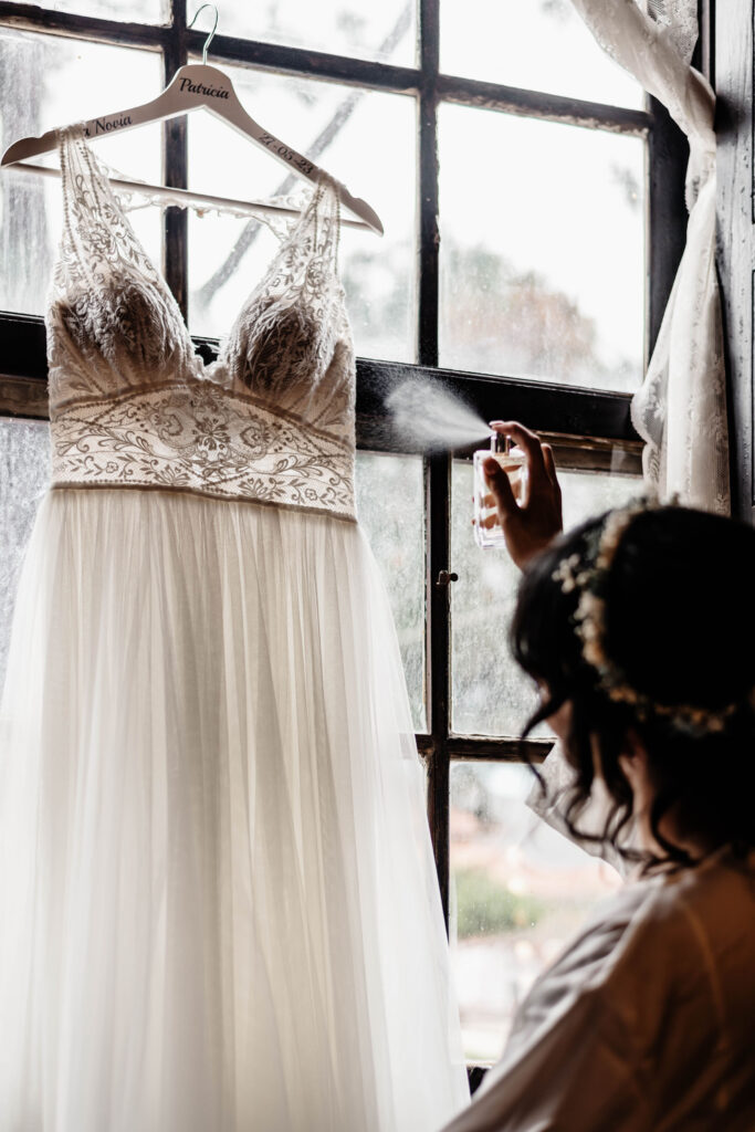 Vestido de novia colgado en una percha de madera frente a una ventana. Una persona está rociando algo sobre el vestido, que tiene detalles de encaje en el corpiño y una falda de tul.