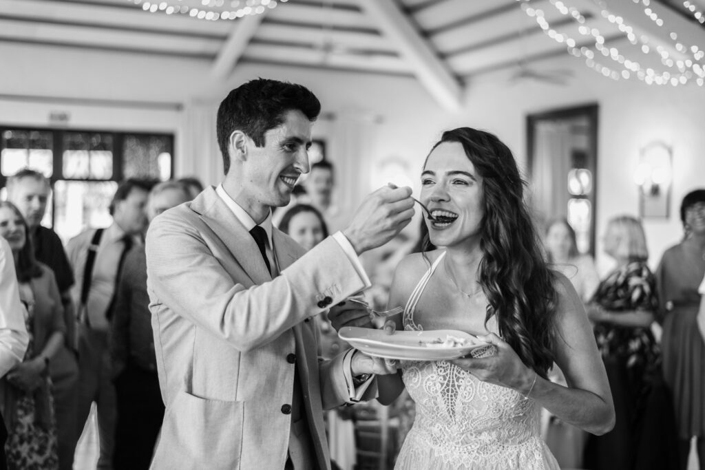 Detalles de boda en blanco y negro: Primer plano de un novio ofreciendo comida a su novia con un vestido de encaje en la recepción, destacando la intimidad y la celebración.