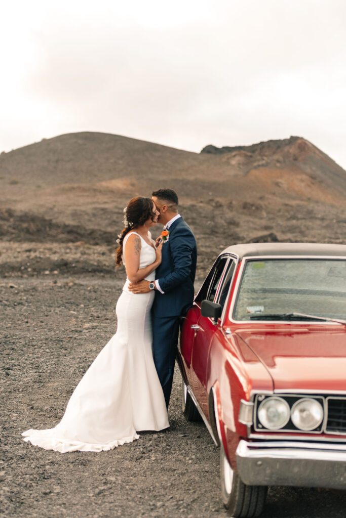 Pareja posando junto a un coche antiguo rojo en un paisaje árido. Ella lleva un vestido blanco de novia con corona de flores, y él viste traje azul oscuro con corbata clara. Fondo montañoso y rocoso que contrasta con la elegancia de la pareja, capturando un momento especial durante una boda