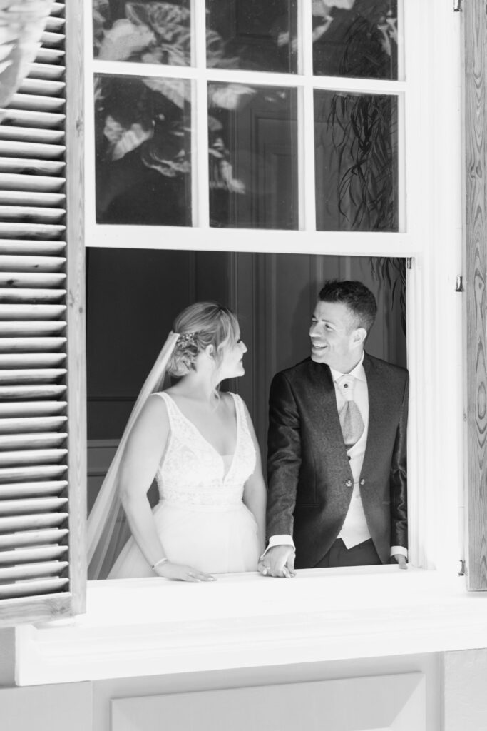 Fotografía de boda: Primer plano de una pareja tomados de la mano, mirando a través de una ventana con contraventanas de madera. La novia lleva un vestido blanco con encaje y velo, y el novio un traje formal