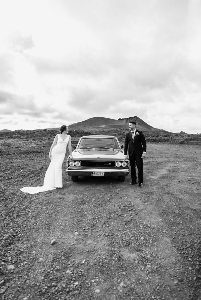 Novia con vestido blanco sin mangas y corona de flores, junto a novio con traje azul oscuro, posando junto a un coche antiguo rojo en un paisaje árido y rocoso con una colina al fondo.