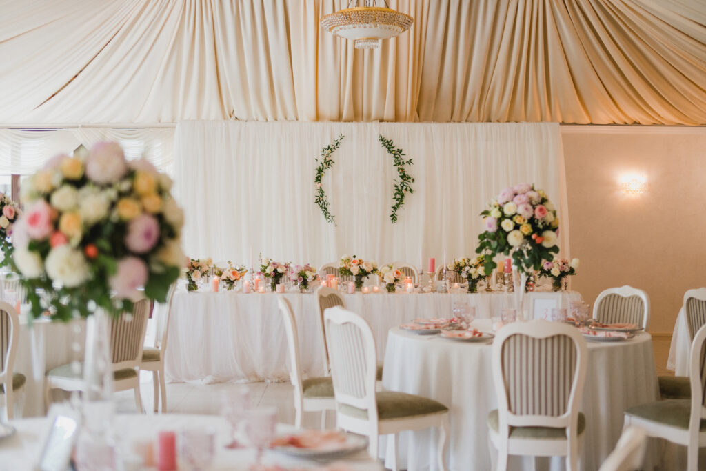 Vista de un salón de bodas con mesas redondas vestidas con manteles blancos, sillas de madera clara y centros de mesa florales, destacando la decoración del fondo con cortinas y guirnaldas verdes, Tendencias bodas 2025.