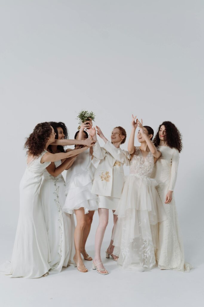 La novia, acompañada por sus damas de honor, todas vestidas de blanco, en un momento de alegría y celebración. La novia levanta un ramo de flores por encima de su cabeza mientras las damas de honor intentan alcanzarlo. La escena captura la tradicional lanzada del ramo en una boda.