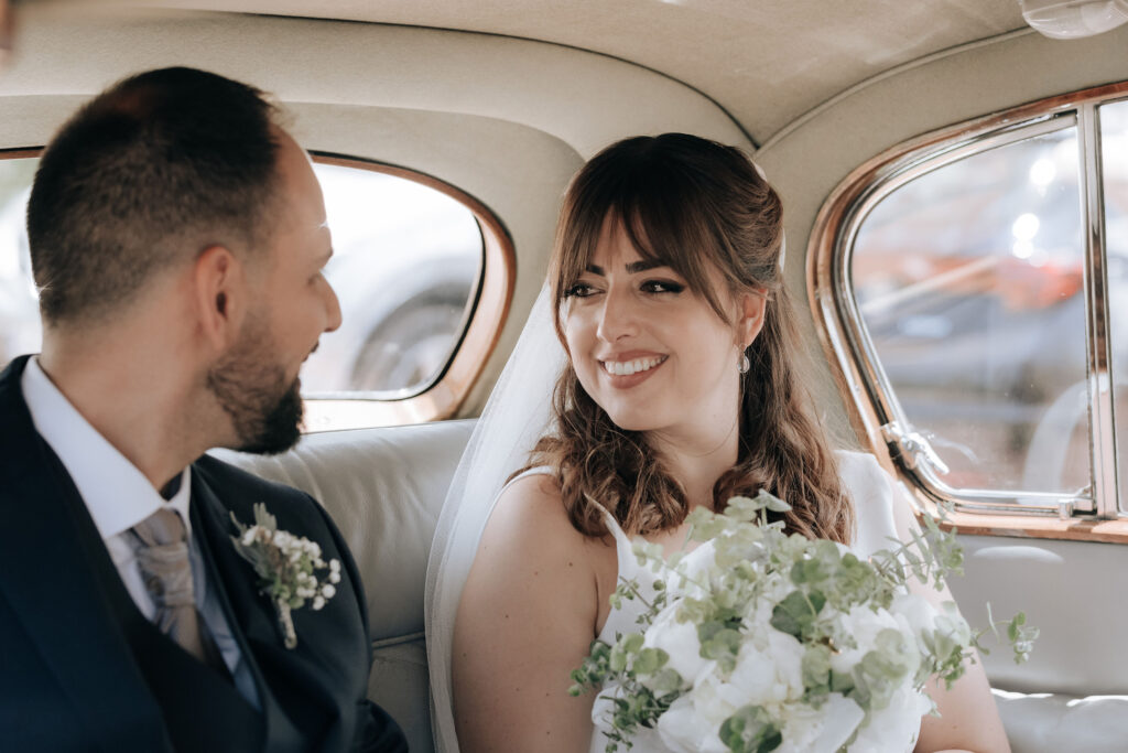 Dos personas vestidas con atuendos formales, una en traje con boutonniere y la otra en un vestido con detalles de plumas. Ambos están junto a un automóvil, frente a una pared lisa y vegetación.