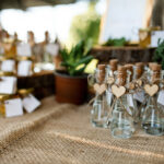 Detalles de boda originales: pequeños frascos de vidrio con tapa de corcho y etiqueta personalizada en forma de corazón, dispuestos sobre una tela de yute rústica.