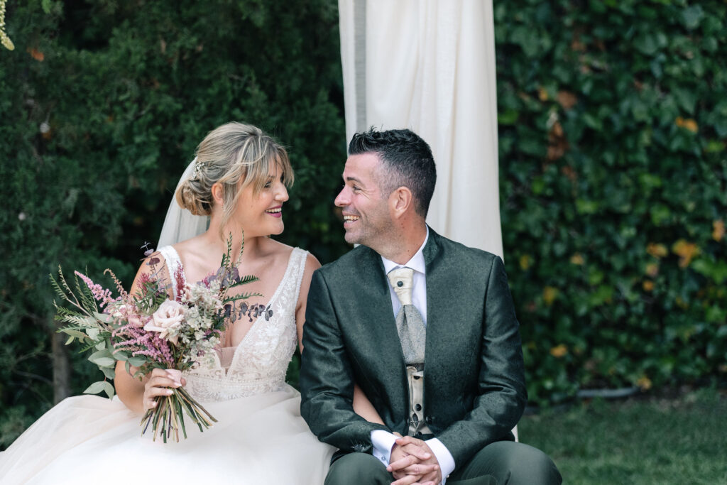 Dos personas sentadas juntas en una boda. La persona a la izquierda lleva un vestido de novia blanco y sostiene un ramo de flores. La persona a la derecha lleva un traje oscuro. Están frente a un fondo de vegetación y una cortina blanca.