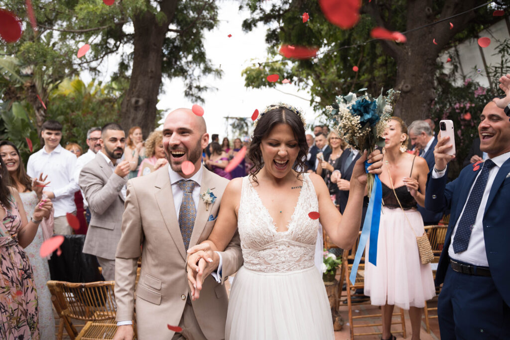 Una pareja camina por el pasillo en su boda, tomados de la mano. Los invitados lanzan pétalos de flores rojas en celebración. La escena es al aire libre con árboles en el fondo. El novio lleva un traje beige y la novia un vestido de encaje blanco.