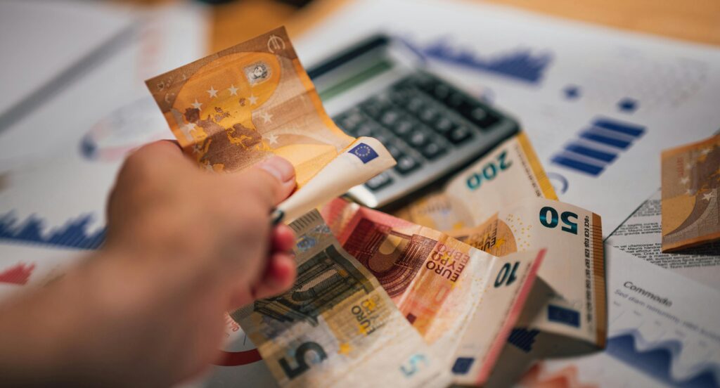 Imagen de una mano contando varios billetes de euro de diferentes denominaciones, incluyendo billetes de 50, 20 y 5 euros. En el fondo, hay una calculadora y documentos financieros con gráficos y diagramas, indicando un contexto financiero o contable. Esta imagen representa una situación común en la gestión financiera o contabilidad, donde el manejo de efectivo y el análisis de datos financieros son tareas esenciales, posiblemente relacionadas con la obtención de una subvención.