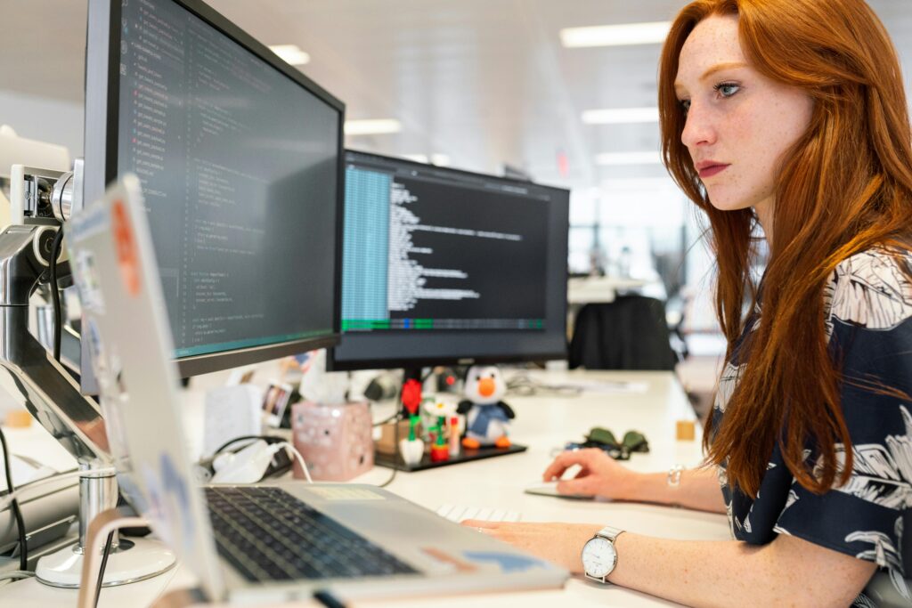 Imagen de una persona trabajando en un escritorio con dos monitores que muestran código. La persona usa un teclado y un ratón, y también hay una laptop en el escritorio. El espacio de trabajo está organizado con varios elementos de oficina. Este entorno moderno refleja una configuración típica para un desarrollador de software, posiblemente relacionado con subvenciones en tecnología.