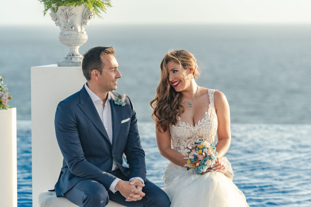 "La imagen muestra a una pareja vestida con trajes formales, probablemente en una boda en Tenerife, con un hermoso paisaje de fondo. La persona de la izquierda lleva un traje azul oscuro y la persona de la derecha un vestido de novia blanco de encaje, sosteniendo un ramo de flores coloridas, y el mar u océano detrás de ellos