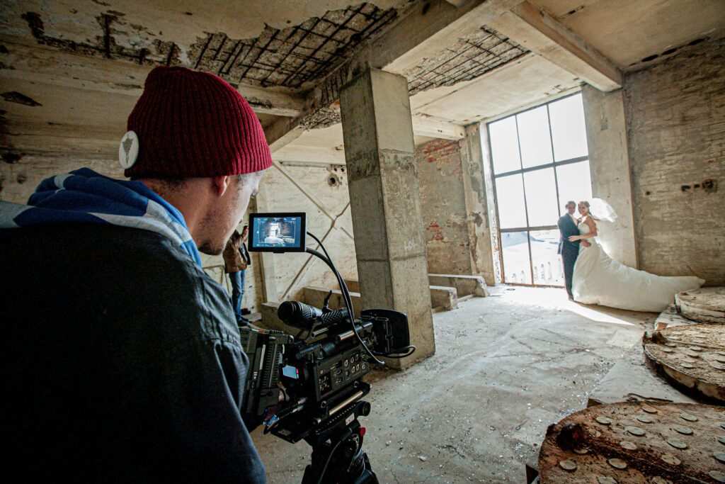 "La imagen muestra una sesión de fotos de bodas en Tenerife, en un edificio industrial abandonado. Un fotógrafo captura a una pareja vestida de boda, con un vestido blanco y un traje oscuro, de pie cerca de una gran ventana en un entorno de concreto y ladrillo.