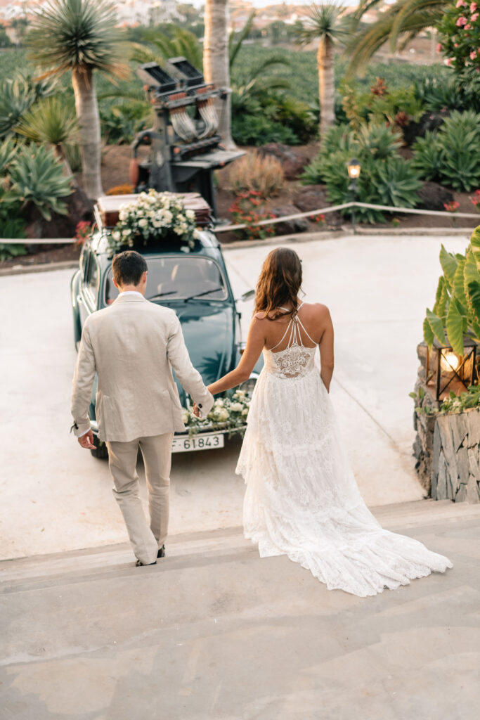 La imagen muestra a una pareja cogidos de la mano y caminando hacia un coche vintage decorado con flores, en el contexto de bodas en Tenerife. La novia lleva un vestido blanco de encaje y el novio un traje claro, con un entorno tropical de exuberante vegetación y palmeras.