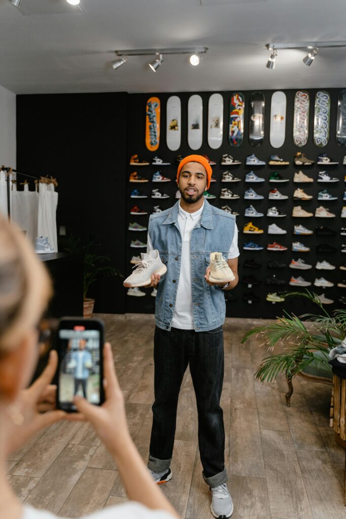 Persona en una tienda de zapatos, sosteniendo un par de zapatillas deportivas en cada mano, mientras es grabada por alguien con un smartphone. El fondo muestra varias zapatillas y tablas de skate en las paredes. Este entorno es ideal para crear videos profesionales en Tenerife.