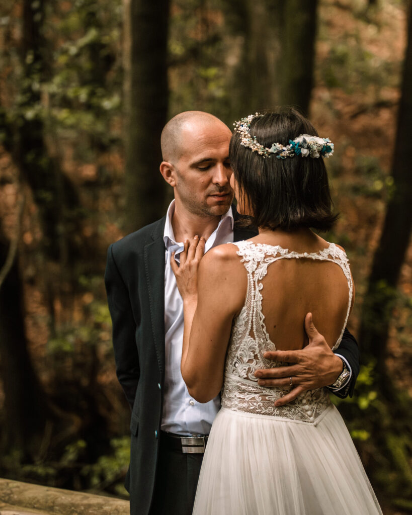 Pareja abrazada durante su boda en un entorno al aire libre con árboles y follaje. La novia lleva un vestido de encaje blanco y una corona de flores, mientras que el novio viste un traje oscuro. Momento íntimo y significativo capturado en la imagen