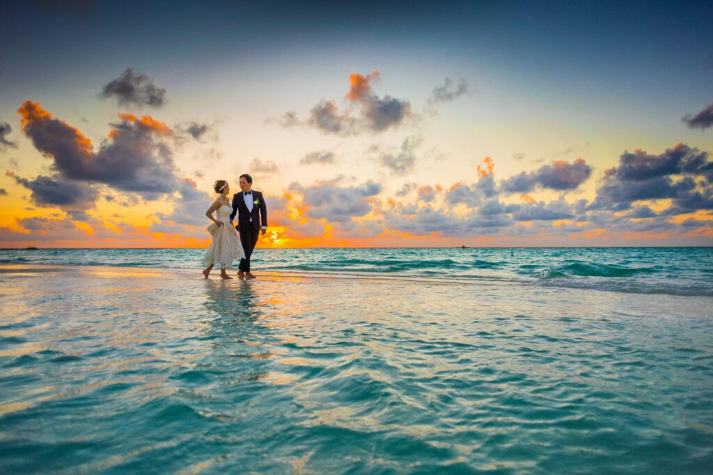 Pareja de recién casados caminando juntos en la orilla del mar durante un atardecer, con un cielo lleno de nubes coloridas reflejándose en el agua