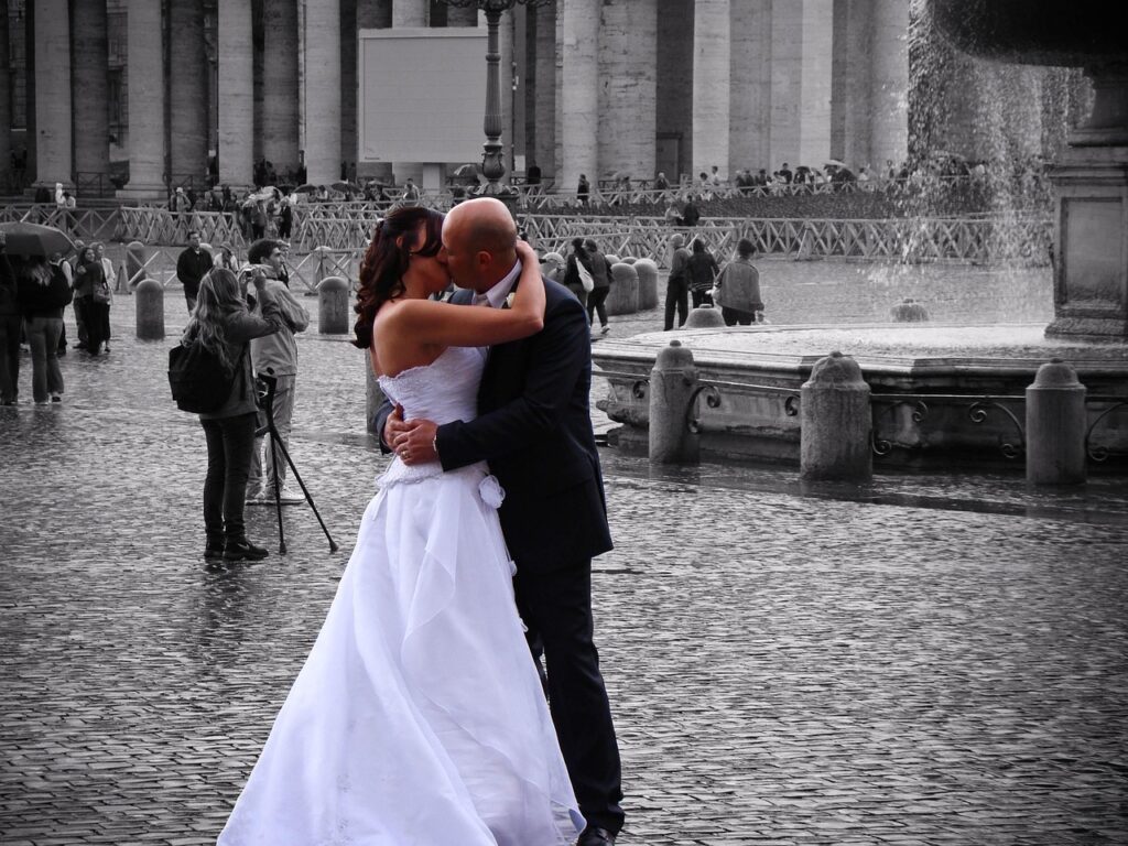 Pareja de recién casados besándose en una plaza pública frente a una fuente grande. La novia lleva un vestido blanco, y el novio, un traje oscuro. Fondo con columnas arquitectónicas y turistas observando