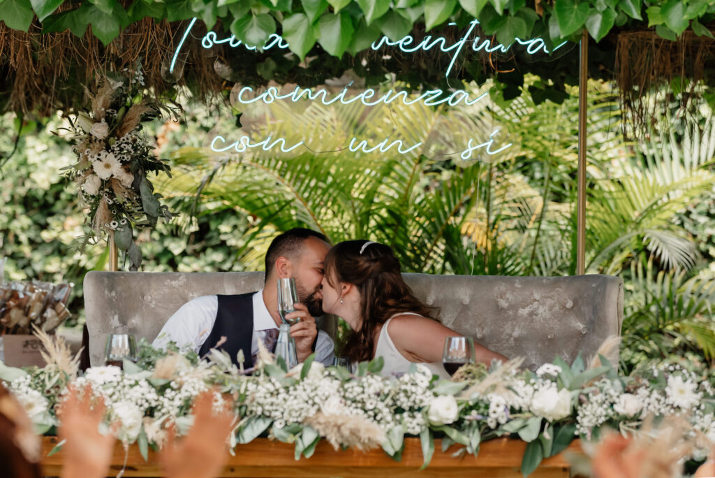 Pareja compartiendo un beso en una mesa decorada con un arreglo floral de flores blancas y vegetación en su boda. Un letrero de neón sobre ellos dice 'Toda aventura comienza con un sí'. Fondo de follaje verde exuberante, creando un ambiente festivo al aire libre.