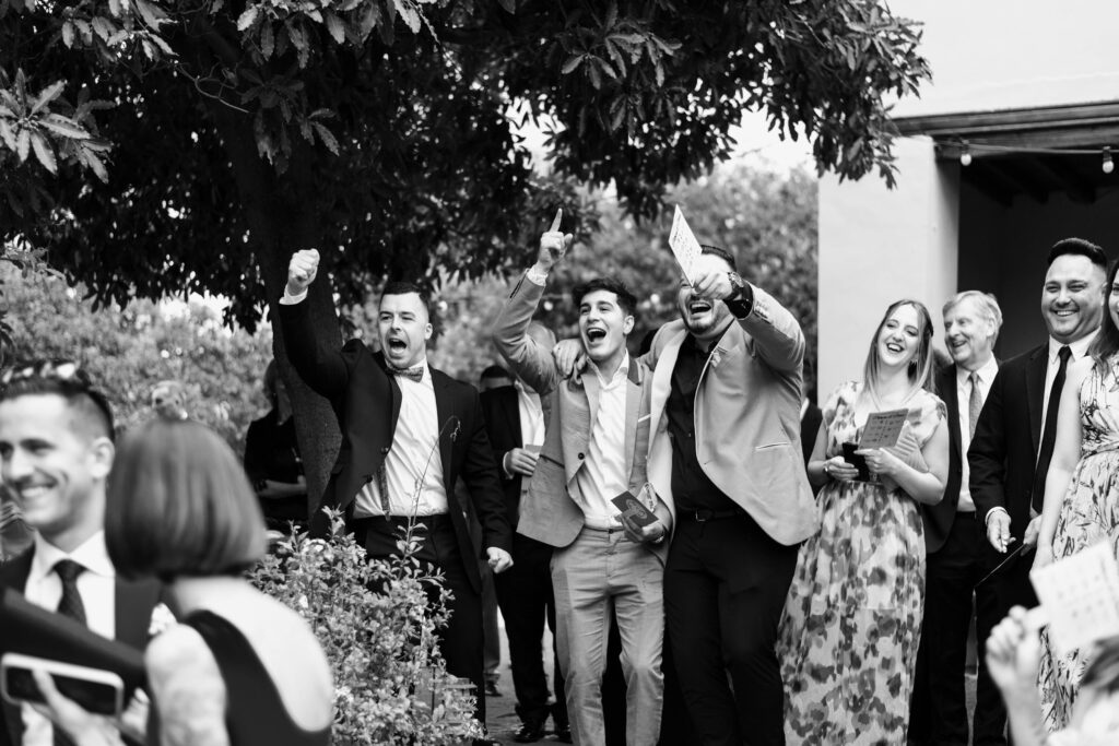 Grupo de personas vestidas de manera formal en un evento al aire libre rodeado de árboles y vegetación. Algunas personas levantan las manos y sostienen bebidas o papeles, creando un ambiente de alegría y celebración