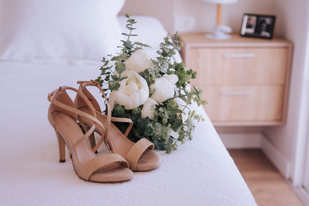 Sandalias beige de tacón alto sobre una cama blanca junto a un ramo de flores blancas con follaje verde. Fondo con mesa de noche de madera, lámpara y marco decorativo en dormitorio minimalista. Preparativos para la fecha de boda.