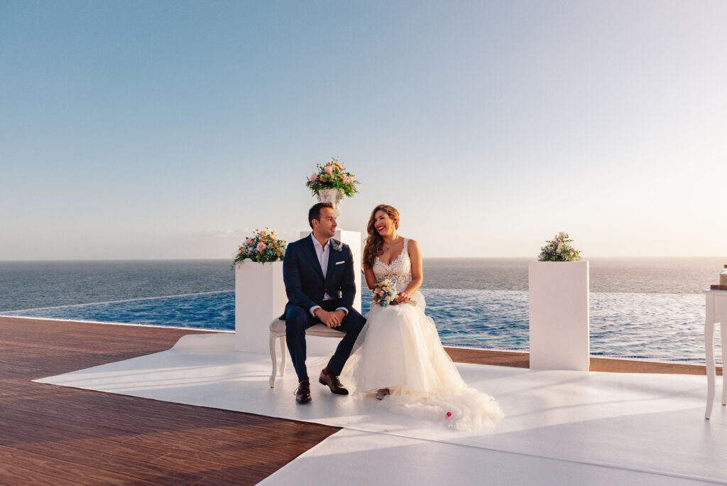 Pareja en su boda, sentados en una plataforma blanca frente al océano. El novio lleva un traje oscuro y la novia un vestido de boda blanco. Decoración con arreglos florales sobre pedestales blancos, con el mar como fondo sereno y escénico.