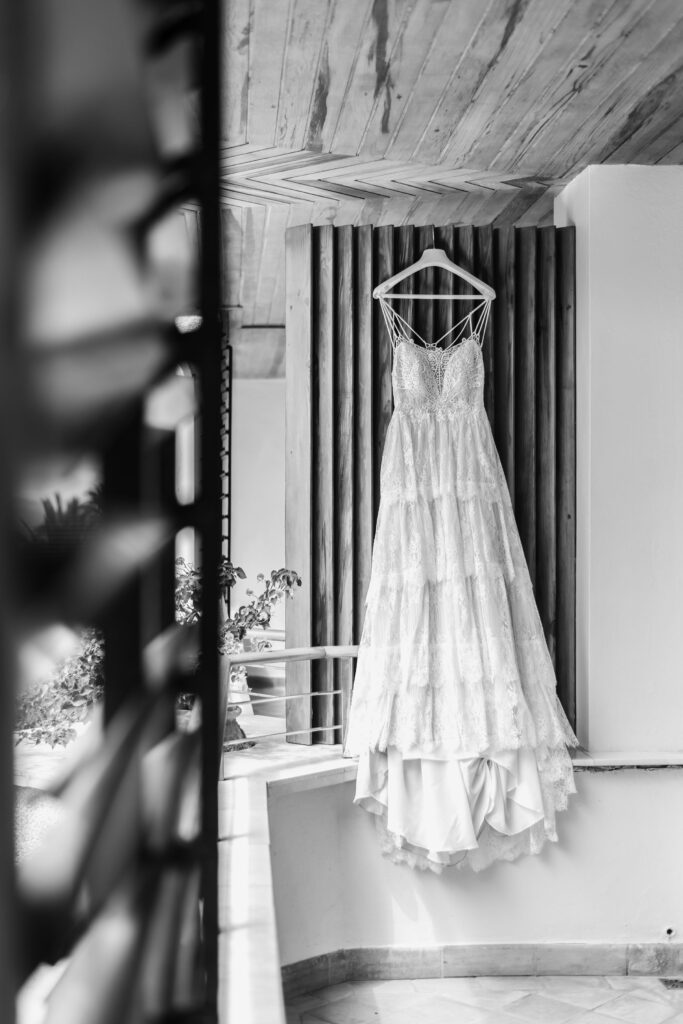 "Vestido traje boda colgado en una percha de madera, sobre una pared con paneles de madera verticales. El vestido es largo, con varias capas de encaje y tirantes finos. La imagen está en blanco y negro, con un techo de madera visible y una barandilla en primer plano.