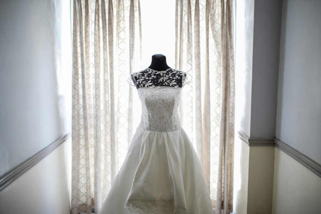 Vestido traje boda blanco exhibido en un maniquí negro. El vestido presenta detalles intrincados de encaje en el corpiño y la parte superior, con patrones florales que se extienden hacia los hombros y el escote. La falda es amplia y parece estar hecha de un tejido liso, similar al satén. En el fondo, hay cortinas claras con un sutil patrón geométrico, y el entorno parece ser un pasillo con paredes de color claro y una moldura decorativa.